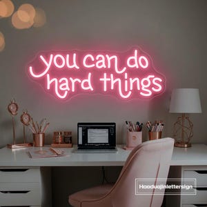 May include: A pink neon sign that reads "you can do hard things" is mounted on a wall above a white desk. The desk is accessorized with rose gold desk supplies, a laptop, and a pink chair. A white lamp with a geometric base sits on the desk.