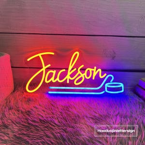May include: A neon sign in yellow and blue, spelling "Jackson" with a hockey stick and puck design. The sign is on a fluffy, pink surface with a wooden background. The text "Hooduojinlettersign" is in the bottom right corner.