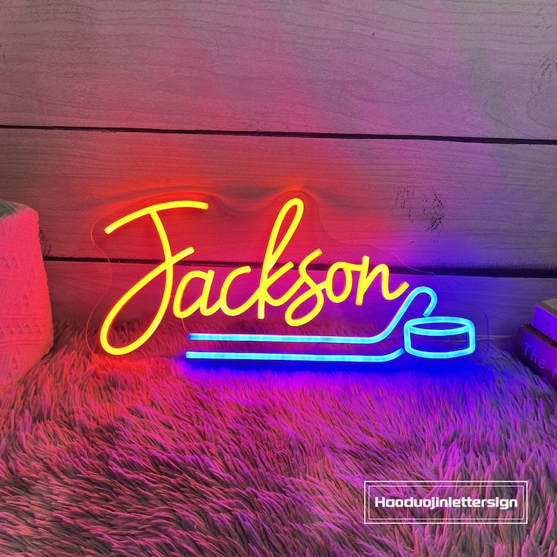 May include: A neon sign in yellow and blue, spelling "Jackson" with a hockey stick and puck design. The sign is on a fluffy, pink surface with a wooden background. The text "Hooduojinlettersign" is in the bottom right corner.