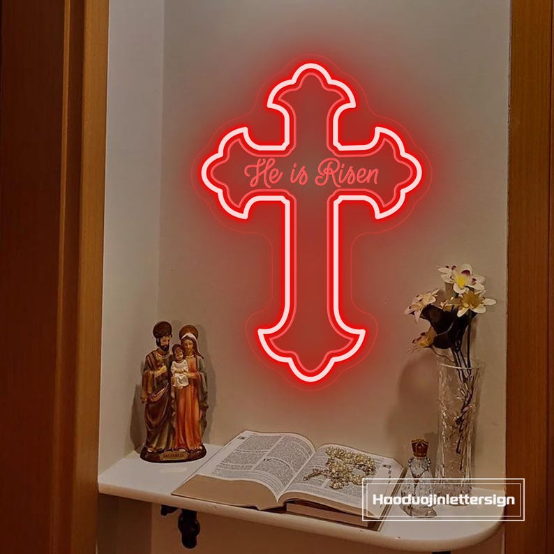 Puede incluir: Un letrero de cruz de ne&oacute;n rojo con las palabras "He is Risen" en un estante blanco. Una estatua de la Sagrada Familia, un libro abierto y un jarr&oacute;n con flores tambi&eacute;n est&aacute;n en el estante.