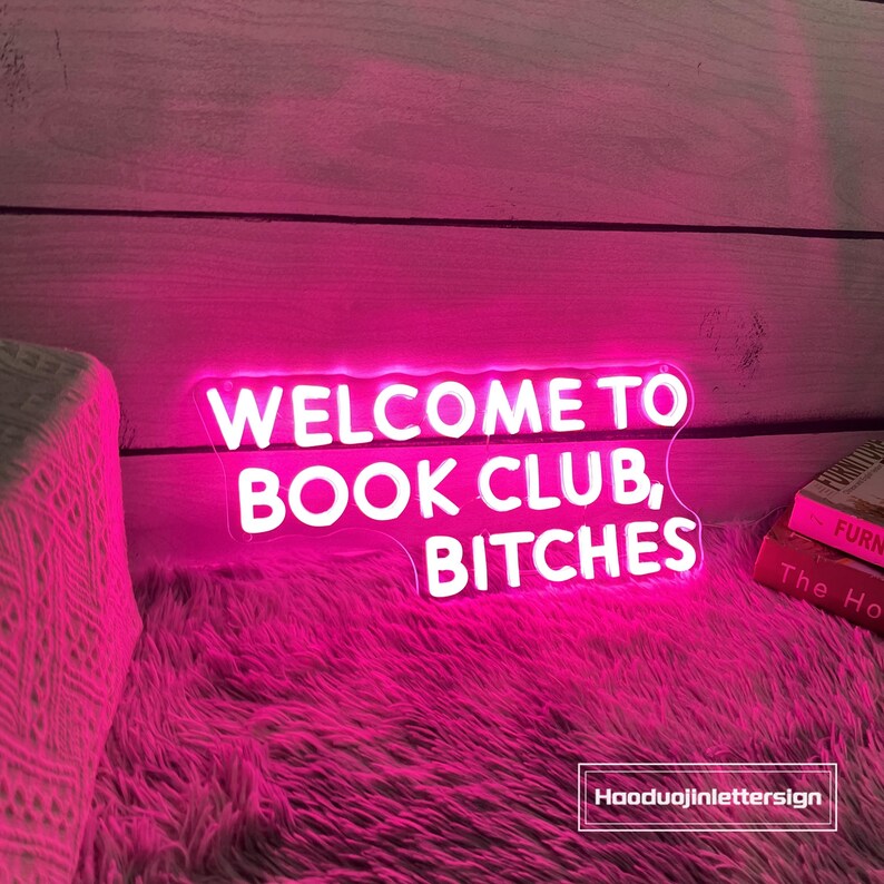 May include: A neon sign in bright pink illuminates the words "WELCOME TO BOOK CLUB, BITCHES" in white. The sign rests on a fluffy pink surface, with a stack of books visible in the background. The sign is a decorative item.