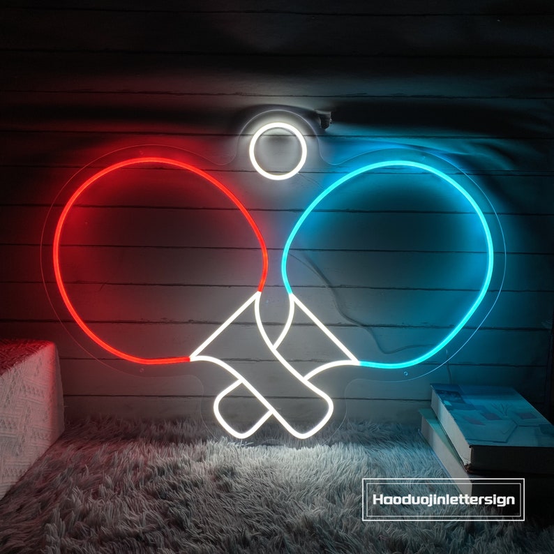 May include: Neon sign depicting two crossed ping pong paddles, one red and one blue, with a white outline. The sign is illuminated and has a white circle at the top.