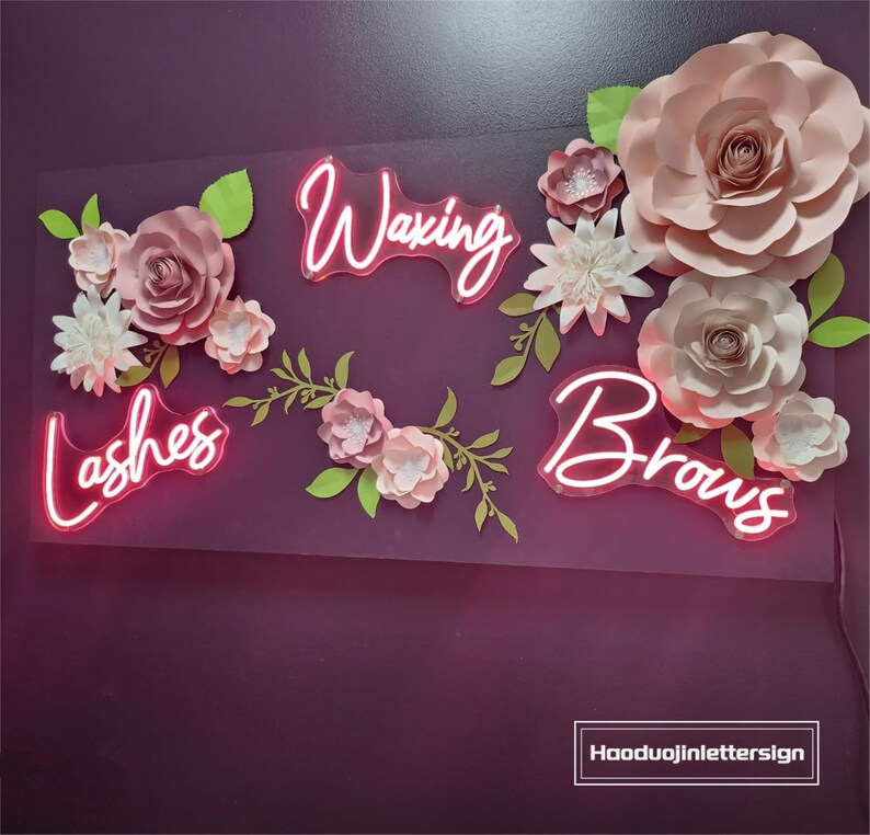 Puede incluir: Flores de papel rosas y vegetaci&oacute;n rodean letreros de ne&oacute;n rosa que dicen "Lashes", "Waxing" y "Brows".