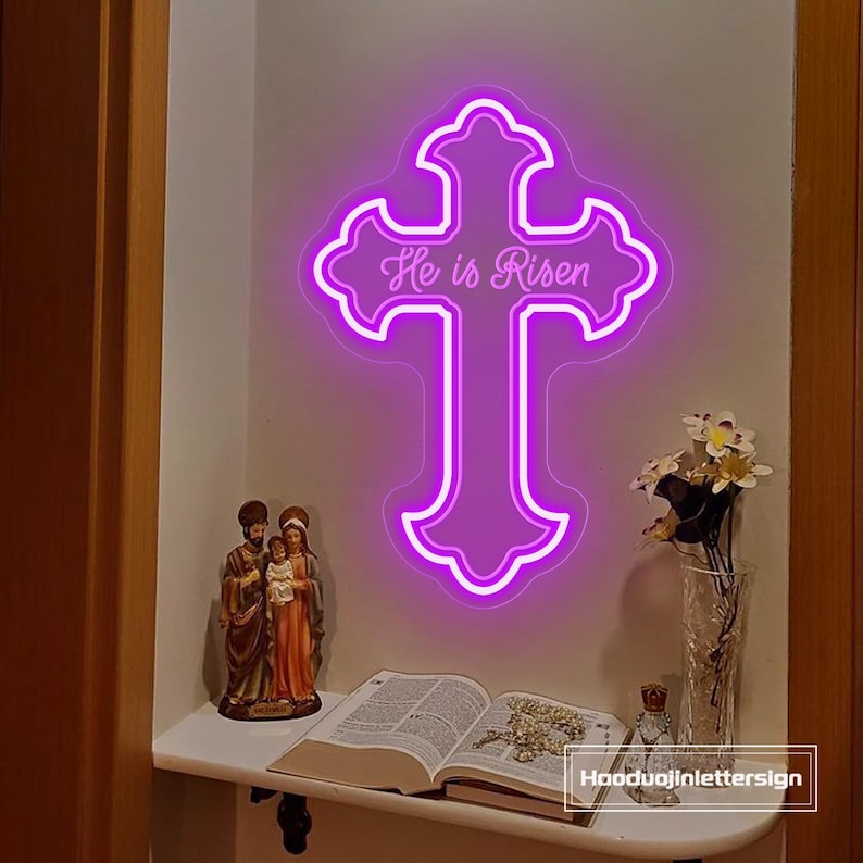 Puede incluir: Una cruz de ne&oacute;n p&uacute;rpura con las palabras "He is Risen". La cruz est&aacute; montada en una pared sobre una estanter&iacute;a con objetos religiosos, incluyendo una estatua, un libro abierto y flores en un jarr&oacute;n.