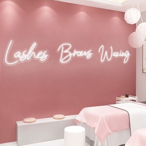 Puede incluir: Una habitaci&oacute;n rosa con un letrero de ne&oacute;n blanco que dice "Lashes Brows Waxing". Hay dos taburetes blancos y una mesa blanca con una manta rosa encima.