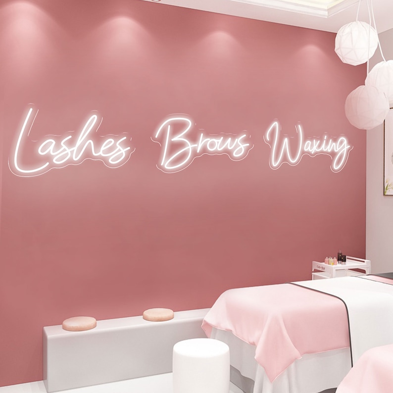 Puede incluir: Una habitaci&oacute;n rosa con un letrero de ne&oacute;n blanco que dice "Lashes Brows Waxing". Hay dos taburetes blancos y una mesa blanca con una manta rosa encima.