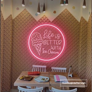 Może przedstawiać: Różowy neon z napisem "Life is Better With Ice Cream" w kawiarni. Znak otoczony jest okręgiem i ilustracją rożka z lodami. Ściany zdobi wzór wafelka, a na zdjęciu widoczne są drewniane stoły.
