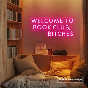 May include: A neon sign in pink illuminates a cozy reading nook. The sign reads "WELCOME TO BOOK CLUB, BITCHES." The room features a built-in bookshelf, a cushioned bench with pillows, and framed artwork.