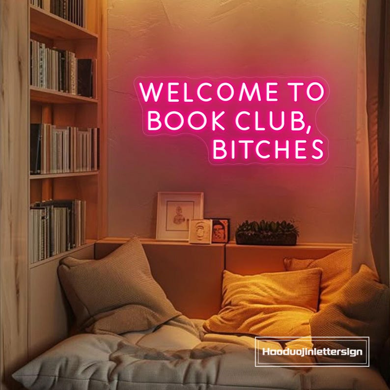 May include: A neon sign in pink illuminates a cozy reading nook. The sign reads "WELCOME TO BOOK CLUB, BITCHES." The room features a built-in bookshelf, a cushioned bench with pillows, and framed artwork.