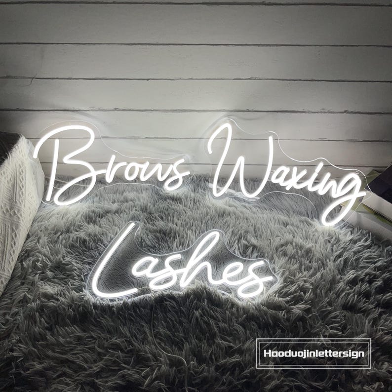 Puede incluir: Letrero de ne&oacute;n blanco con las palabras "Brows Waxing Lashes" en una fuente cursiva. El letrero est&aacute; sobre una superficie blanca y esponjosa.