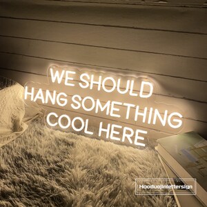 May include: A neon sign with the text "WE SHOULD HANG SOMETHING COOL HERE" in white. The sign is on a clear backing and is placed on a fluffy, white surface. The sign is lit up and the text is easy to read.
