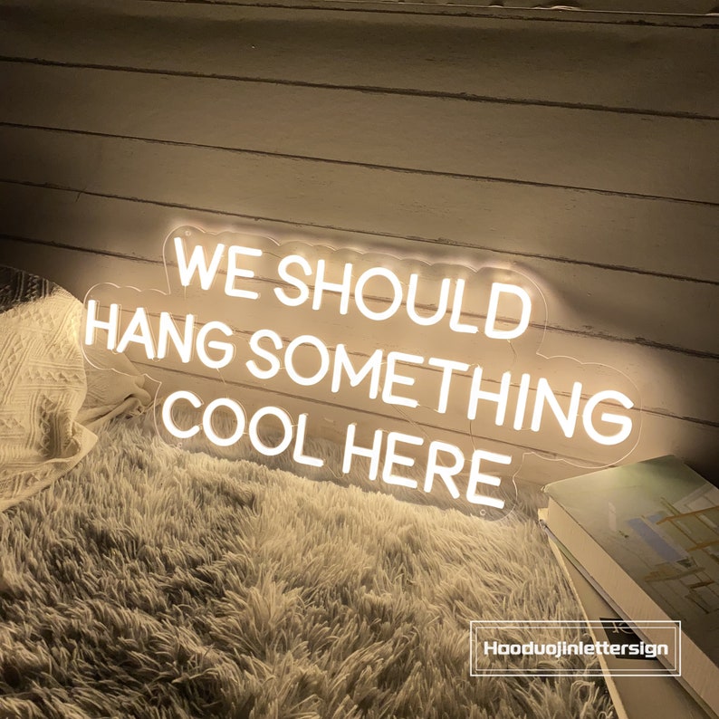 May include: A neon sign with the text "WE SHOULD HANG SOMETHING COOL HERE" in white. The sign is on a clear backing and is placed on a fluffy, white surface. The sign is lit up and the text is easy to read.