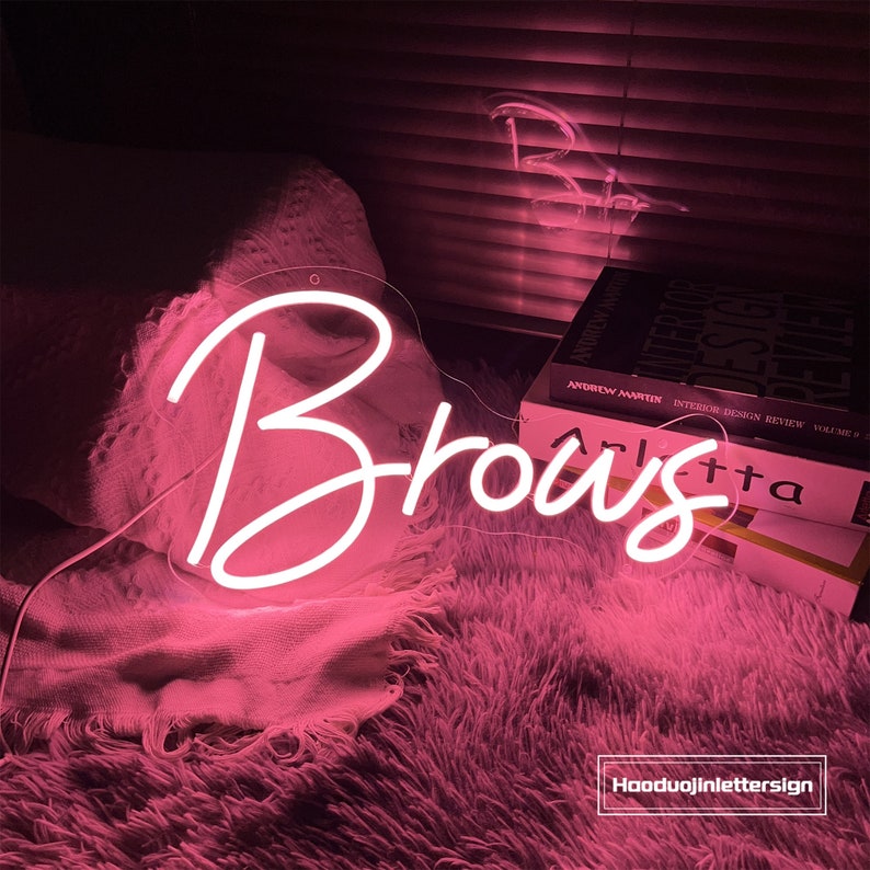 Puede incluir: Un letrero de ne&oacute;n rosa con la palabra "Brous" en una fuente cursiva. El letrero est&aacute; sobre una alfombra rosa peluda con libros en el fondo.