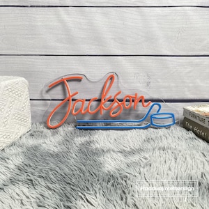 May include: A neon sign with the name "Jackson" in orange script, accompanied by a blue hockey stick and puck design. The sign is mounted on a clear backing and rests on a gray, fluffy surface.