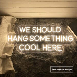 May include: A white neon sign with the text "WE SHOULD HANG SOMETHING COOL HERE". The sign is on a gray, fluffy surface. The sign is lit up and has a warm glow.