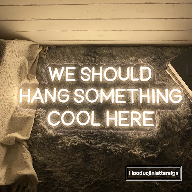May include: A white neon sign with the text "WE SHOULD HANG SOMETHING COOL HERE". The sign is on a gray, fluffy surface. The sign is lit up and has a warm glow.