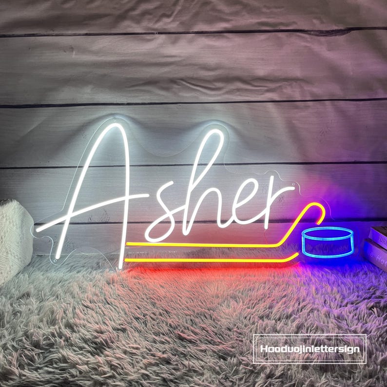 May include: A white neon sign spelling "Asher" with a hockey puck design. The sign has a red and yellow base with a blue and white hockey puck. The sign is on a gray rug with a wooden background.