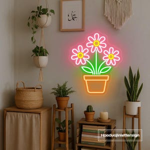 May include: A neon sign in the shape of a flower pot with three flowers. The flowers are pink with yellow centers and green stems and leaves. The sign is mounted on a wall with other decorative items, including plants and a woven basket.