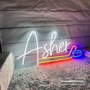 May include: A neon sign displaying the name "Asher" in white script. The sign includes a hockey puck graphic and is outlined with red, yellow, and blue neon lights. The sign is placed on a white wooden background.