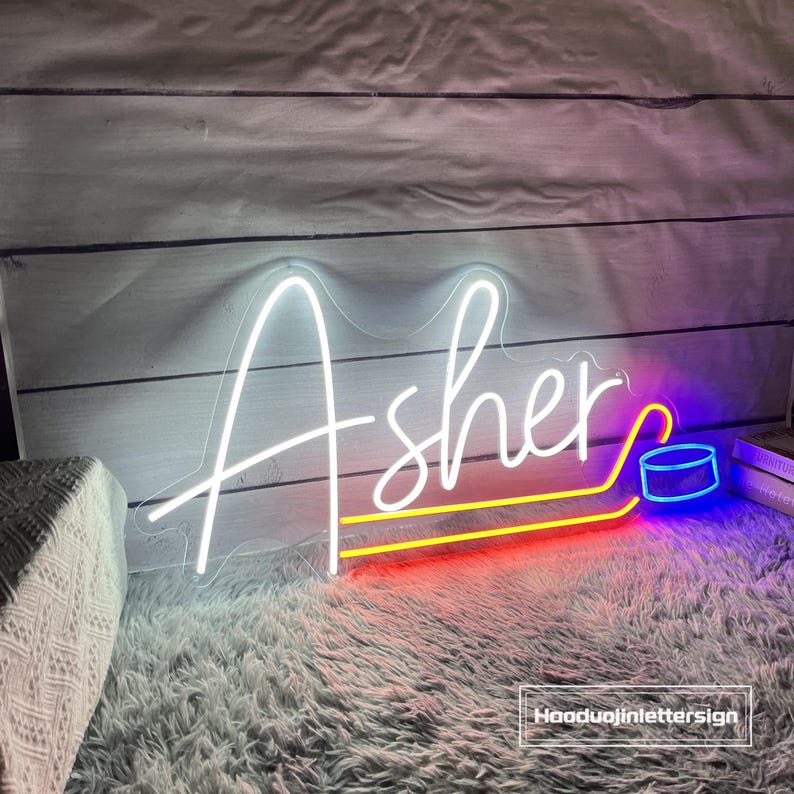 May include: A neon sign displaying the name "Asher" in white script. The sign includes a hockey puck graphic and is outlined with red, yellow, and blue neon lights. The sign is placed on a white wooden background.