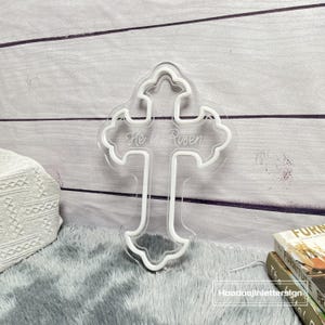 Puede incluir: Una cruz de acr&iacute;lico transparente con un contorno blanco y las palabras "He is Risen" grabadas en el centro. La cruz tiene un borde decorativo festoneado. La cruz se muestra sobre un fondo claro de vetas de madera.