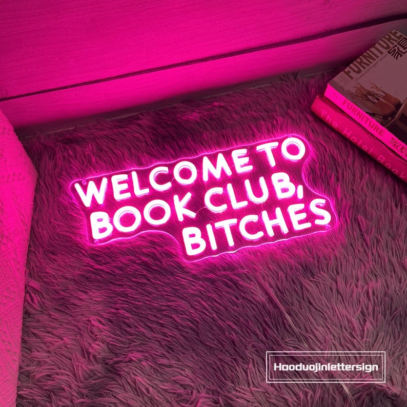 May include: A neon sign in a pink hue reads "WELCOME TO BOOK CLUB, BITCHES" in white letters. The sign is placed on a fluffy, pinkish-gray rug. Two books are visible in the background.