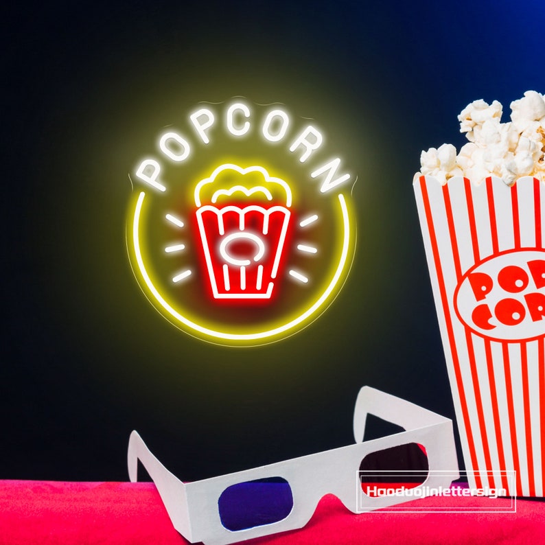 May include: A yellow neon sign with the word "POPCORN" in white letters and a red and white popcorn bucket icon. The sign is illuminated and has a glow effect. There is a red and white striped popcorn box and 3D glasses in the foreground.