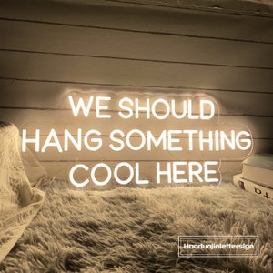 May include: A neon sign with the text "WE SHOULD HANG SOMETHING COOL HERE" in a white, glowing font. The sign is mounted on a wall and sits above a fluffy, white rug. The sign is a decorative item.