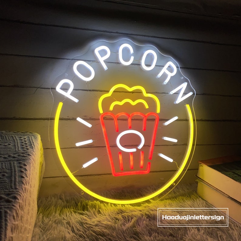 May include: A white neon sign with the word "POPCORN" in a circle with a yellow outline. Inside the circle is a red and yellow neon image of a popcorn bucket.