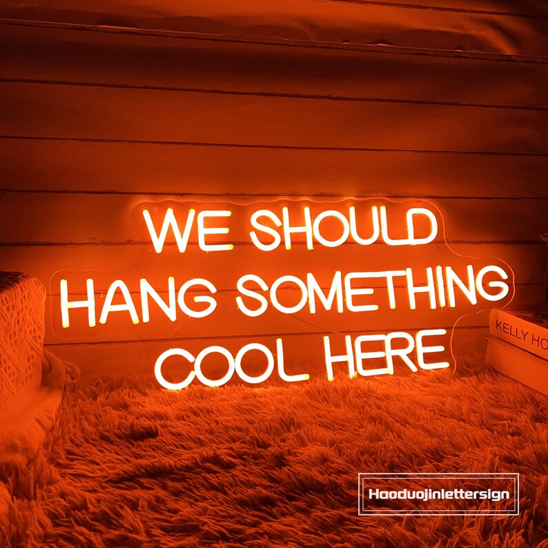 May include: An orange neon sign with the text "WE SHOULD HANG SOMETHING COOL HERE". The sign is illuminated against a wooden background and a fluffy orange surface. The sign is a decorative item.