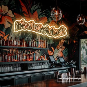 Może przedstawiać: Wnętrze baru z neonowym szyldem z napisem "it's shoto'clock". Szyld znajduje się nad półkami wypełnionymi butelkami i szklankami. Tłem jest tropikalny mural kwiatowy. Bar ma nowoczesny design.