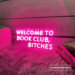 May include: A neon sign with the text "WELCOME TO BOOK CLUB, BITCHES" in white against a pink background. The sign is placed on a fluffy pink surface with a wooden backdrop. Books are visible.