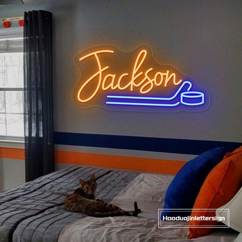 May include: A neon sign in the shape of the name "Jackson" with a hockey puck design. The sign is illuminated in orange and blue. The sign is mounted on a gray wall above a bed with pillows.