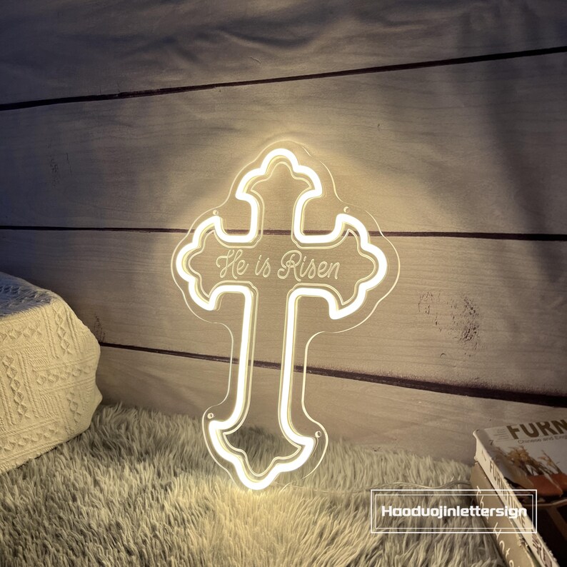 Puede incluir: Un letrero LED transparente en forma de cruz de acr&iacute;lico con las palabras "He is Risen". El letrero tiene un contorno blanco brillante y est&aacute; iluminado. El letrero est&aacute; sobre un fondo de madera.