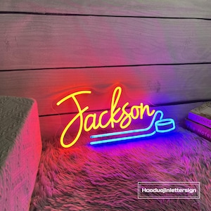 May include: A neon sign in the name "Jackson" with a hockey puck design. The word "Jackson" is in yellow, with a red and blue gradient. The sign is on a fluffy, pink surface.