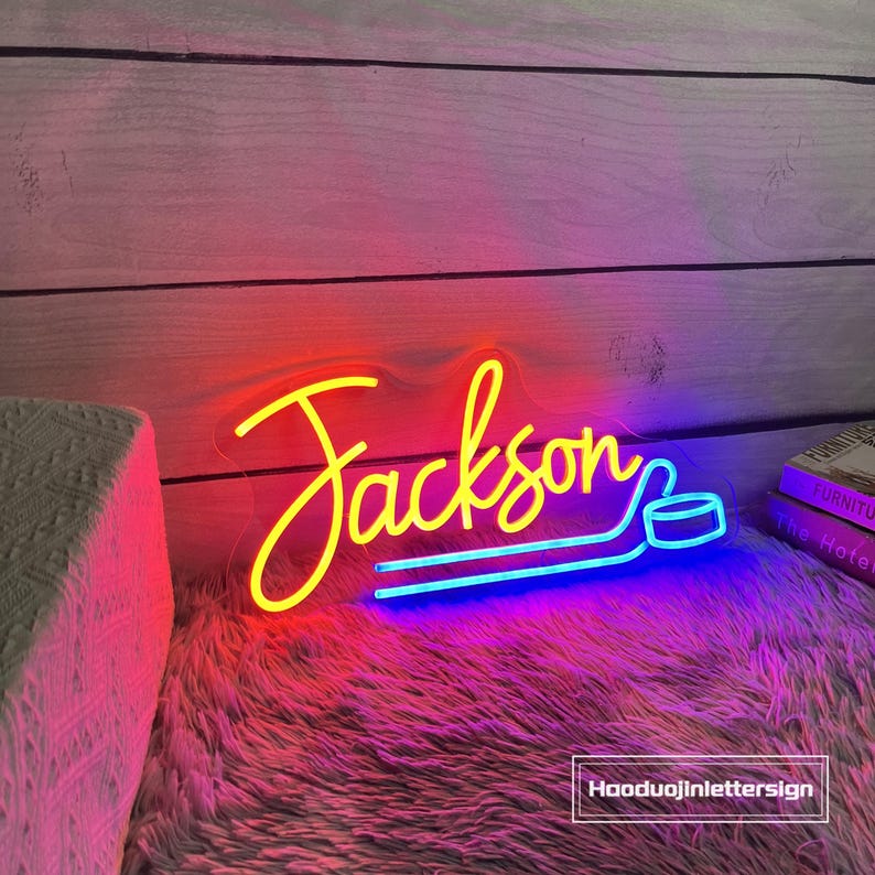 May include: A neon sign in the name "Jackson" with a hockey puck design. The word "Jackson" is in yellow, with a red and blue gradient. The sign is on a fluffy, pink surface.