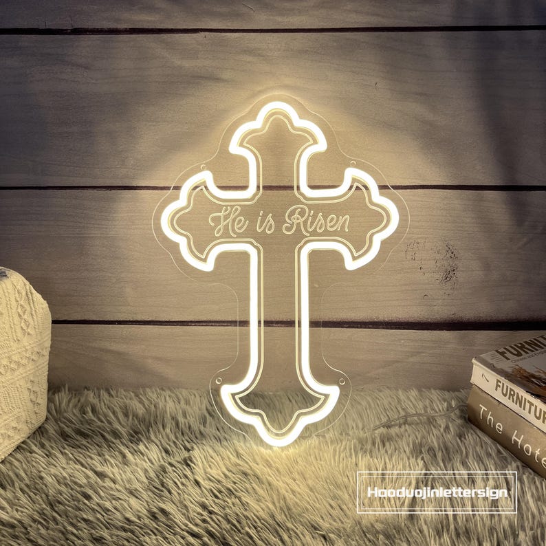 Puede incluir: Una cruz de ne&oacute;n con la frase "He is Risen" en escritura cursiva. La cruz se ilumina con una luz blanca c&aacute;lida y est&aacute; montada sobre un respaldo transparente. El fondo es una pared de madera.