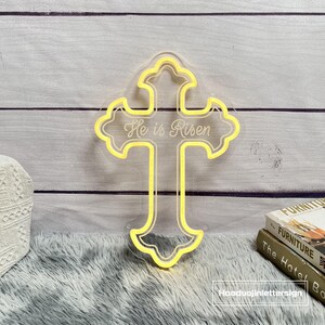 Puede incluir: Una cruz amarilla ne&oacute;n con el texto "He is Risen" en una fuente de escritura. La cruz est&aacute; delineada en un material acr&iacute;lico transparente. El fondo es una superficie de madera gris claro.