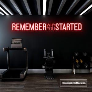 Könnte beinhalten: Ein Heim-Fitnessstudio mit Laufband, Trainingsgeräten und Hanteln. Ein rotes Neon-Schild an der schwarzen Wand zeigt "REMEMBER WHY YOU STARTED". Der Boden ist holzähnlich, und die Decke hat weiße und schwarze Balken.