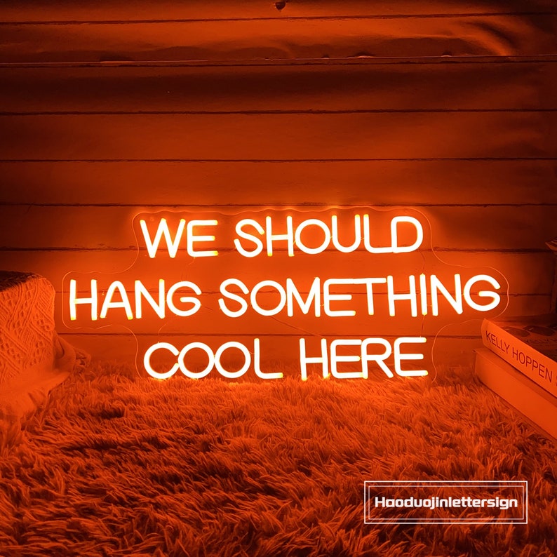 May include: An orange neon sign with the text "WE SHOULD HANG SOMETHING COOL HERE" in a white, sans-serif font. The sign is mounted on an orange wooden wall and sits above a fluffy orange rug. The sign's brand name is "Hooduojinlettersign."