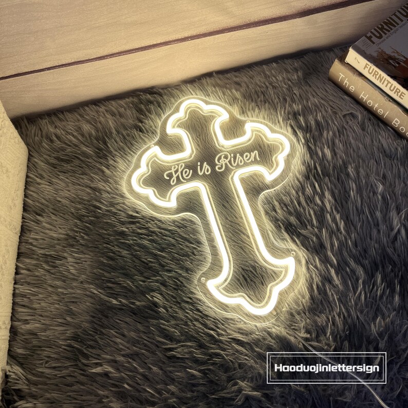 Puede incluir: Una cruz de ne&oacute;n brillante con la frase "He is Risen" escrita. La cruz est&aacute; delineada con una luz blanca c&aacute;lida y se encuentra sobre una superficie gris texturizada. Decoraci&oacute;n religiosa.