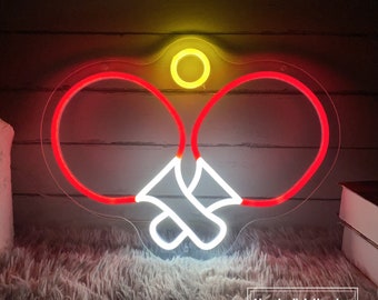 May include: A neon sign depicting two red ping pong paddles and a white ball. The sign is mounted on a clear acrylic backing. The sign is lit up and glowing.