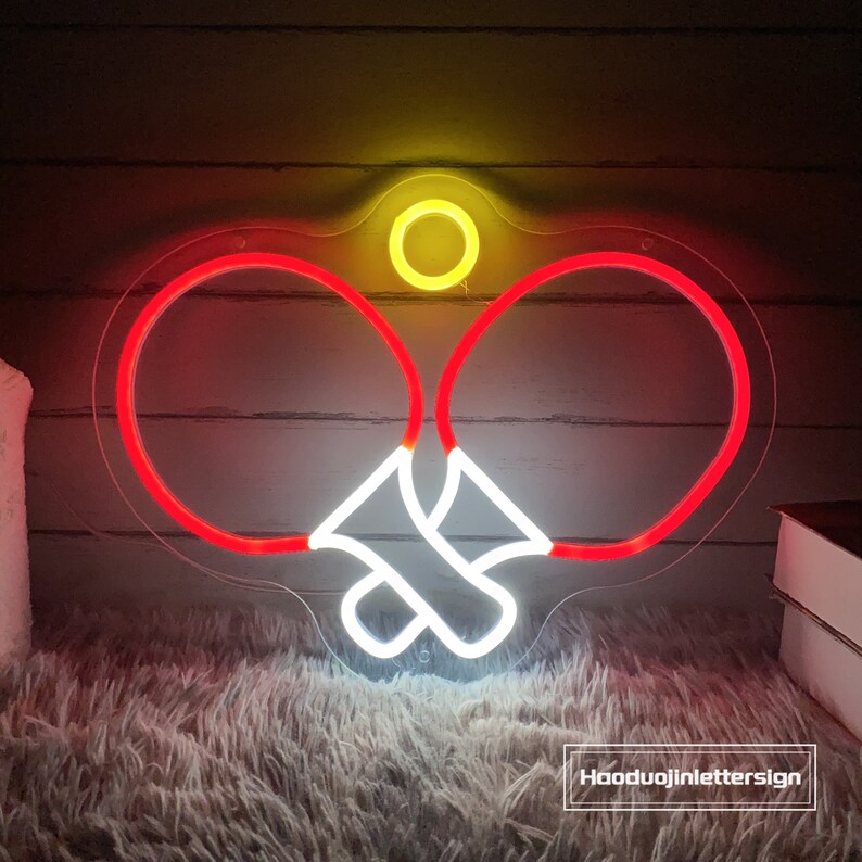 May include: A neon sign depicting two red ping pong paddles and a white ball. The sign is mounted on a clear acrylic backing. The sign is lit up and glowing.