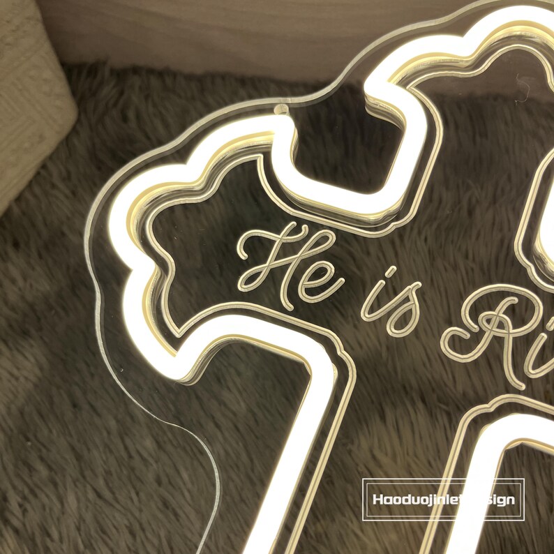 Puede incluir: Un letrero de acr&iacute;lico transparente con las palabras "He is Risen" en fuente cursiva, iluminado por una luz LED blanca c&aacute;lida. El letrero tiene un borde decorativo y est&aacute; sobre un fondo suave y texturizado.