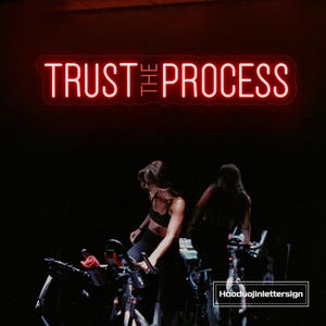 Op de afbeelding: Een rood neonbord met de tekst "TRUST THE PROCESS" staat boven twee mensen op hometrainers. Het bord bevindt zich in een donkere kamer en de mensen dragen sportkleding.