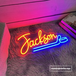 May include: A neon sign with the name "Jackson" in yellow script, with a blue hockey stick design. The sign is on a fluffy gray surface, with a pink and purple background. The text "Haoduojinlettersign" is in the bottom right corner.