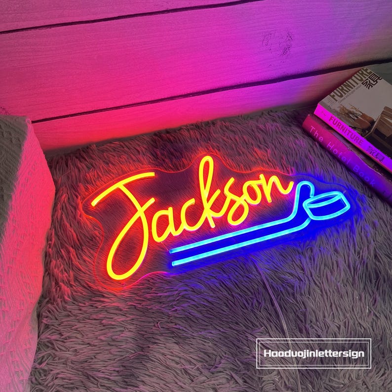 May include: A neon sign with the name "Jackson" in yellow script, with a blue hockey stick design. The sign is on a fluffy gray surface, with a pink and purple background. The text "Haoduojinlettersign" is in the bottom right corner.