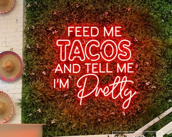 May include: Neon sign with the text "Feed me tacos and tell me I'm pretty" on a green wall with a white brick wall in the background.