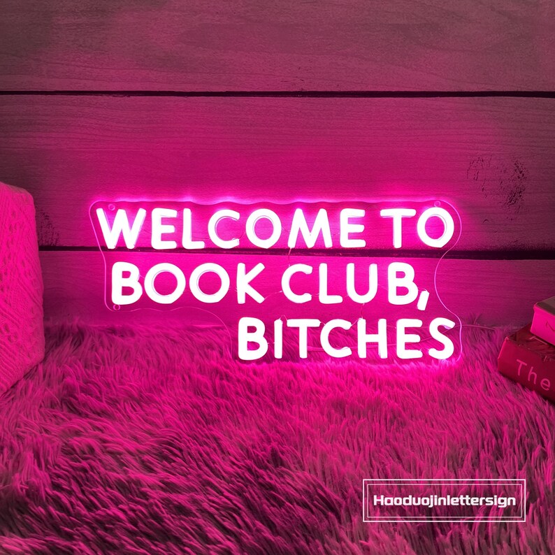 May include: A neon pink sign with the words "WELCOME TO BOOK CLUB, BITCHES" in white lettering. The sign is on a pink fluffy surface, with a wooden background. The sign is a decorative item.