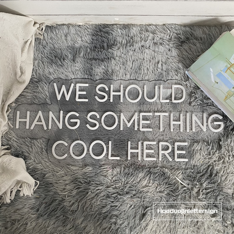 May include: A clear acrylic sign with the text "WE SHOULD HANG SOMETHING COOL HERE" in white, lit-up letters. The sign is on a gray, fluffy surface, with a white blanket and a book visible in the background.